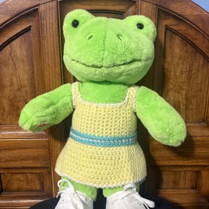 Puede incluir: Una rana de peluche verde con un vestido amarillo de ganchillo con un cinturón azul y blanco. La rana tiene zapatillas blancas. El fondo es una puerta de madera.