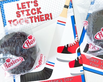 May include: A Valentine's Day card featuring a hockey stick and a pair of ice skates. The card reads "Let's Stick Together!" and "Happy Valentine's Day! Your friend, Logan". The card is decorated in red, white and blue. Two chocolate Hostess cupcakes are included.