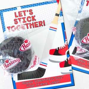Könnte beinhalten: Eine Valentinstagskarte mit einem Hockeyschläger und einem Paar Schlittschuhen. Die Karte enthält den Text "Let's Stick Together!" und "Happy Valentine's Day! Your friend, Logan". Die Karte ist in den Farben Rot, Weiß und Blau verziert. Zwei Schokoladen-Hostess-Cupcakes sind enthalten.