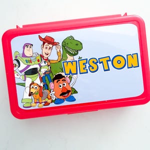 May include: A rectangular red plastic lunch box featuring a light blue lid with a graphic print of Toy Story characters including Buzz Lightyear, Woody, Rex, Slinky Dog, and Mr. Potato Head, with the name WESTON printed in bold yellow letters.