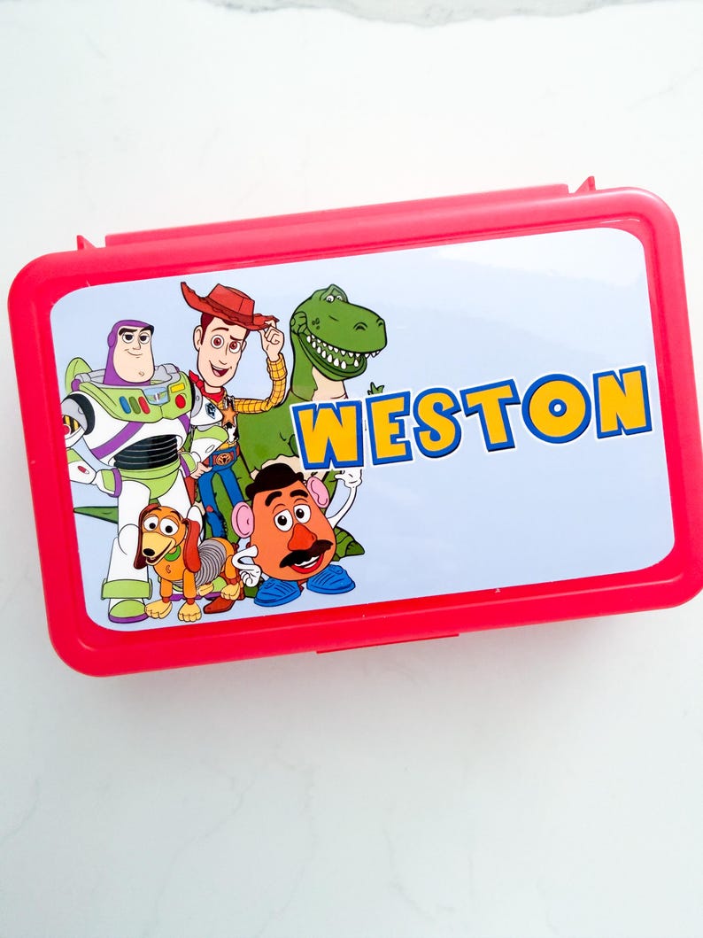 May include: A rectangular red plastic lunch box featuring a light blue lid with a graphic print of Toy Story characters including Buzz Lightyear, Woody, Rex, Slinky Dog, and Mr. Potato Head, with the name WESTON printed in bold yellow letters.