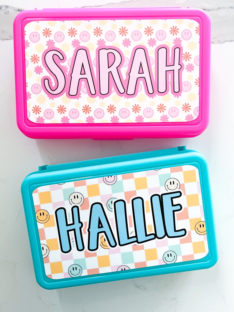 K&ouml;nnte beinhalten: Zwei rechteckige Brotdosen aus Kunststoff mit Deckeln. Die obere Dose ist leuchtend pink mit einem wei&szlig;en Etikett, das rosa Smileys, orangefarbene Blumen und den Namen SARAH in gro&szlig;en schwarzen Buchstaben zeigt. Die untere Dose ist t&uuml;rkis mit einem Karomuster.