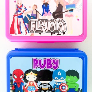May include: A pink rectangular lunch box featuring a graphic of five figures and the name FLYNN, and a blue rectangular lunch box featuring a graphic of five superheroes and the name RUBY. Both containers have hinged lids and colorful printed labels.