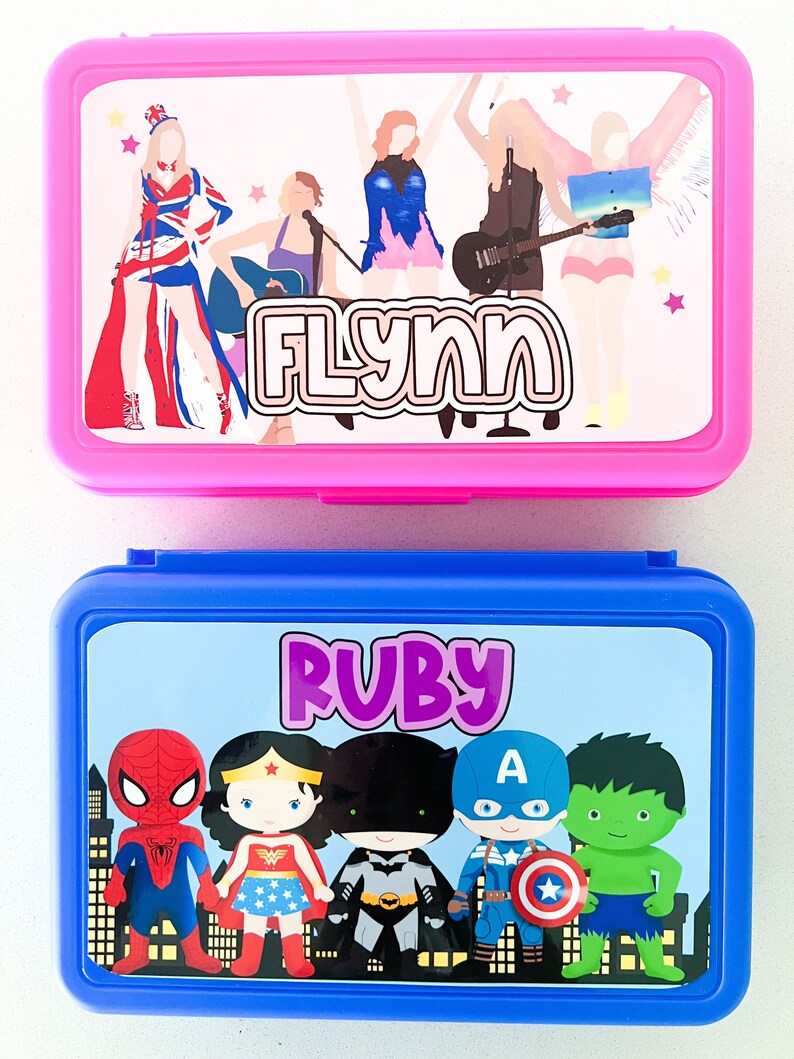 May include: A pink rectangular lunch box featuring a graphic of five figures and the name FLYNN, and a blue rectangular lunch box featuring a graphic of five superheroes and the name RUBY. Both containers have hinged lids and colorful printed labels.