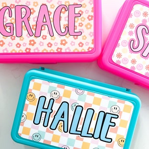 K&ouml;nnte beinhalten: Zwei rechteckige Kunststoff-Federm&auml;ppchen in leuchtendem Pink und T&uuml;rkis. Das pinke M&auml;ppchen zeigt den Namen GRACE in pinken Buchstaben auf einem Blumenmuster. Das t&uuml;rkise M&auml;ppchen zeigt den Namen HALLIE in blauen Buchstaben auf einem Karomuster.