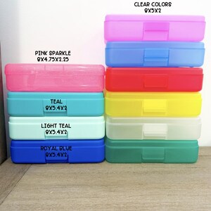 May include: A collection of rectangular plastic pencil boxes in various colors including pink sparkle, teal, light teal, royal blue, and clear shades. Each box features a hinged lid and a latch, with printed labels indicating dimensions in inches.
