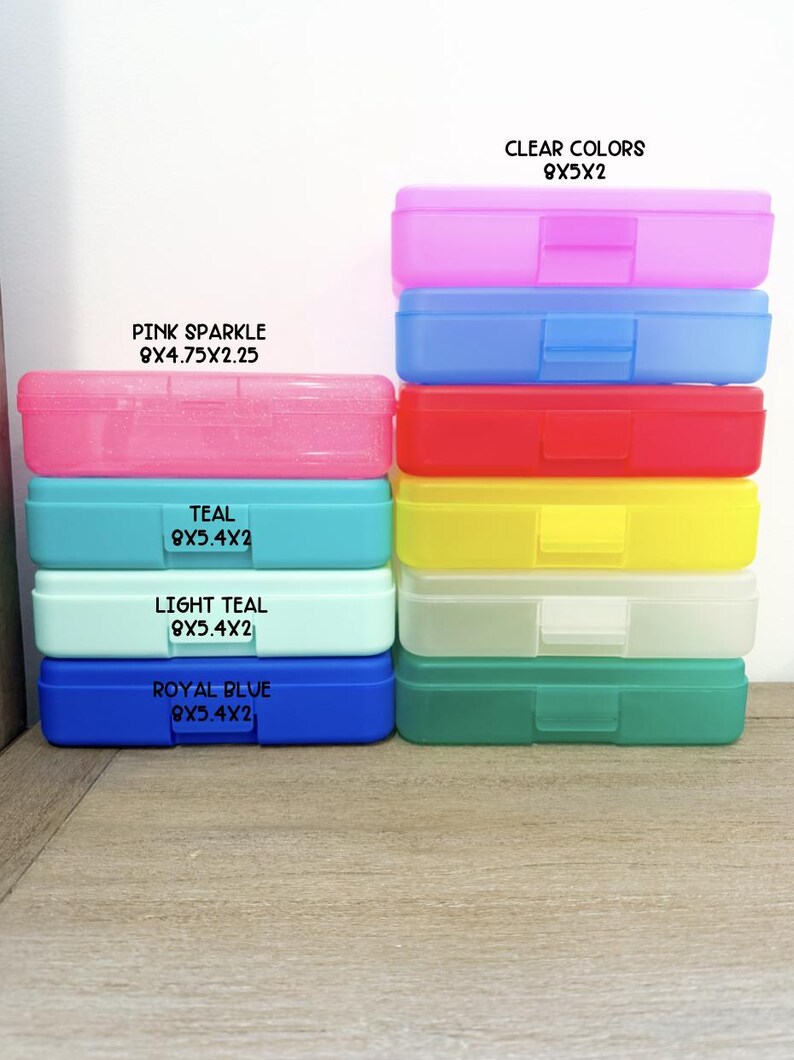 May include: A collection of rectangular plastic pencil boxes in various colors including pink sparkle, teal, light teal, royal blue, and clear shades. Each box features a hinged lid and a latch, with printed labels indicating dimensions in inches.
