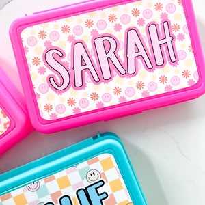 K&ouml;nnte beinhalten: Eine leuchtend pinke, rechteckige Brotdose mit dem Namen SARAH in gro&szlig;en schwarzen Buchstaben auf wei&szlig;em Grund, verziert mit kleinen rosa Smileys und orangefarbenen Blumen. Darunter befindet sich eine t&uuml;rkisfarbene Brotdose mit dem Namen HALLIE.