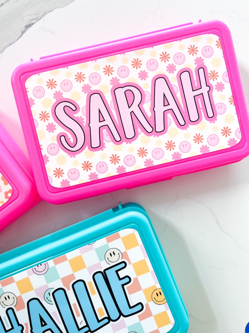 K&ouml;nnte beinhalten: Eine leuchtend pinke, rechteckige Brotdose mit dem Namen SARAH in gro&szlig;en schwarzen Buchstaben auf wei&szlig;em Grund, verziert mit kleinen rosa Smileys und orangefarbenen Blumen. Darunter befindet sich eine t&uuml;rkisfarbene Brotdose mit dem Namen HALLIE.