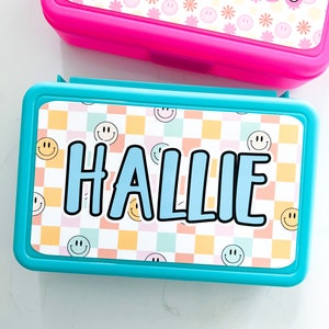 K&ouml;nnte beinhalten: Eine rechteckige t&uuml;rkisfarbene Brotdose mit einem personalisierten Etikett, das den Namen HALLIE in fetten blauen Buchstaben zeigt. Der Hintergrund des Etiketts zeigt ein pastellfarbenes Karomuster mit kleinen gelben und blauen Smileys.