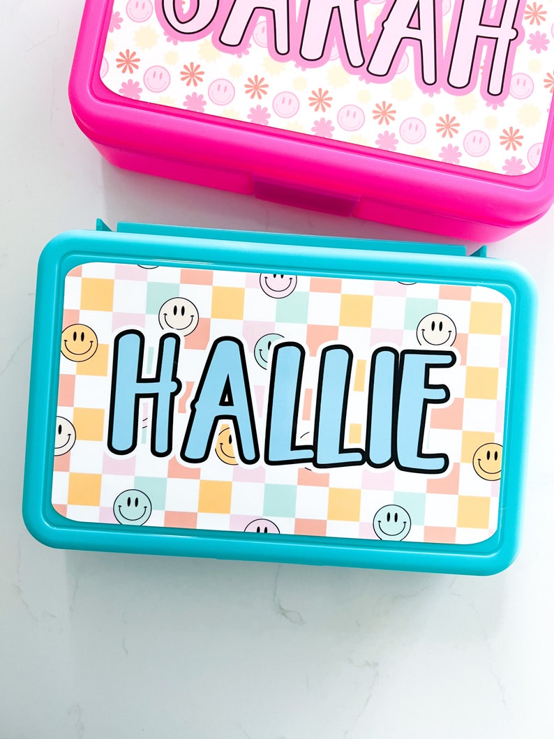 K&ouml;nnte beinhalten: Eine rechteckige t&uuml;rkisfarbene Brotdose mit einem personalisierten Etikett, das den Namen HALLIE in fetten blauen Buchstaben zeigt. Der Hintergrund des Etiketts zeigt ein pastellfarbenes Karomuster mit kleinen gelben und blauen Smileys.