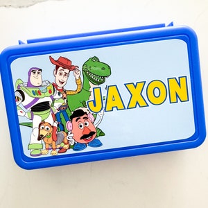 May include: A rectangular blue plastic lunch box featuring a light blue lid with a graphic print of Toy Story characters including Buzz Lightyear, Woody, Rex, Slinky Dog, and Mr. Potato Head, with the name JAXON printed in large yellow letters.