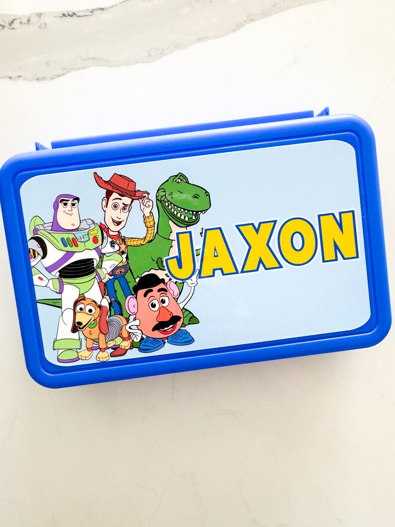 May include: A rectangular blue plastic lunch box featuring a light blue lid with a graphic print of Toy Story characters including Buzz Lightyear, Woody, Rex, Slinky Dog, and Mr. Potato Head, with the name JAXON printed in large yellow letters.