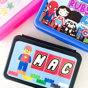 May include: Three rectangular lunch boxes in pink, blue, and black. The pink box has glittery text and stars. The blue box features superhero characters and the name Ruby. The black box displays a block-style figure and the name MAC.