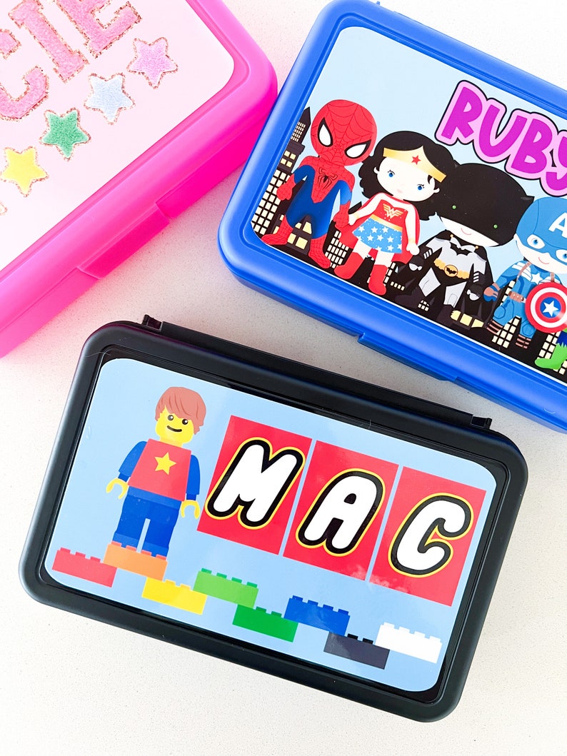 May include: Three rectangular lunch boxes in pink, blue, and black. The pink box has glittery text and stars. The blue box features superhero characters and the name Ruby. The black box displays a block-style figure and the name MAC.
