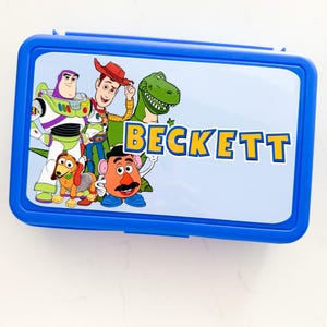 May include: A rectangular blue plastic lunch box featuring a light blue lid with a graphic print of Toy Story characters including Buzz Lightyear, Woody, Rex, Slinky Dog, and Mr. Potato Head, with the name BECKETT printed in yellow and blue letters.