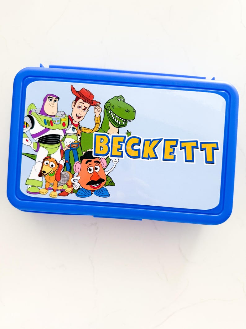 May include: A rectangular blue plastic lunch box featuring a light blue lid with a graphic print of Toy Story characters including Buzz Lightyear, Woody, Rex, Slinky Dog, and Mr. Potato Head, with the name BECKETT printed in yellow and blue letters.