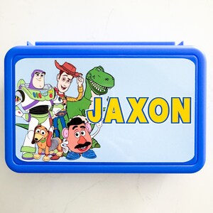 May include: A rectangular blue plastic lunch box featuring a light blue lid with a graphic print of Toy Story characters including Woody, Buzz Lightyear, Rex, Slinky Dog, and Mr. Potato Head, with the name JAXON printed in large yellow letters.