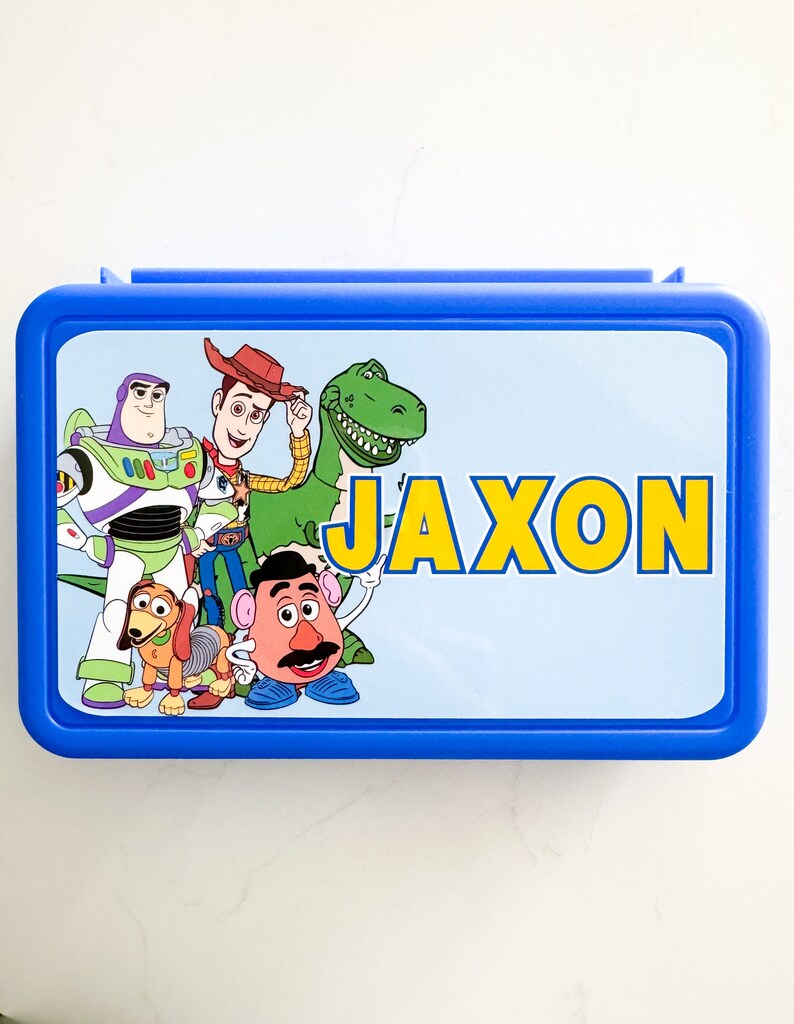 May include: A rectangular blue plastic lunch box featuring a light blue lid with a graphic print of Toy Story characters including Woody, Buzz Lightyear, Rex, Slinky Dog, and Mr. Potato Head, with the name JAXON printed in large yellow letters.