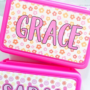 K&ouml;nnte beinhalten: Zwei rechteckige, leuchtend pinke Kunststoffbeh&auml;lter mit personalisierten Aufklebern. Der obere Beh&auml;lter zeigt den Namen GRACE in pinken Buchstaben auf wei&szlig;em Hintergrund mit orangen und pinken Blumen. Der untere Beh&auml;lter zeigt SARAH.