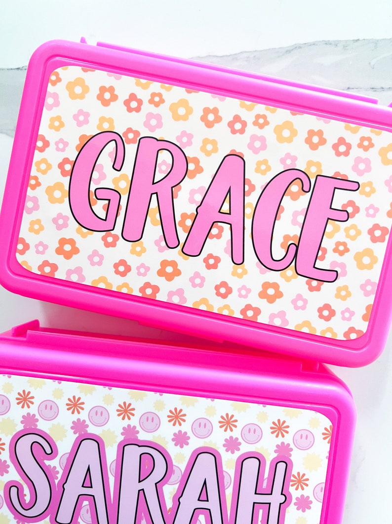 K&ouml;nnte beinhalten: Zwei rechteckige, leuchtend pinke Kunststoffbeh&auml;lter mit personalisierten Aufklebern. Der obere Beh&auml;lter zeigt den Namen GRACE in pinken Buchstaben auf wei&szlig;em Hintergrund mit orangen und pinken Blumen. Der untere Beh&auml;lter zeigt SARAH.