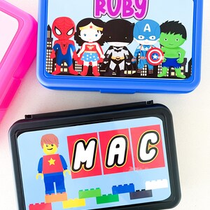 May include: A blue rectangular lunch box featuring a graphic of superheroes and the name Ruby, and a black rectangular lunch box featuring a Lego-style character, the name MAC, and colorful building blocks printed on the lids.