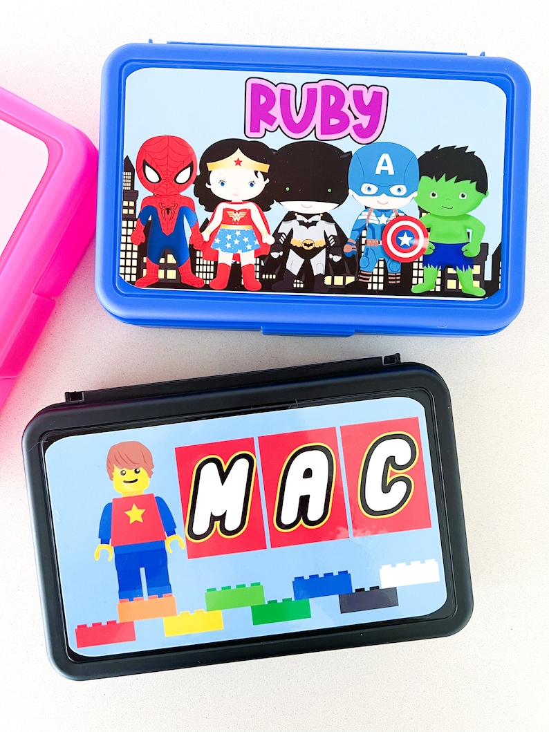 May include: A blue rectangular lunch box featuring a graphic of superheroes and the name Ruby, and a black rectangular lunch box featuring a Lego-style character, the name MAC, and colorful building blocks printed on the lids.