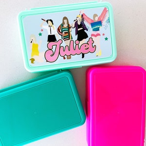 May include: Three rectangular plastic lunch boxes in mint green, teal, and hot pink. The mint green box features a graphic illustration of five figures and the name Juliet in pink script lettering, accented by small green and red star shapes.