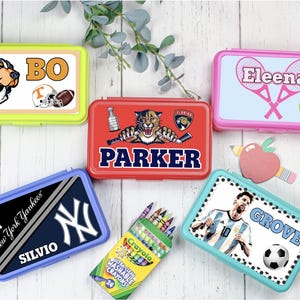 May include: Five rectangular plastic pencil cases in lime green, red, dark blue, pink, and light blue, each featuring personalized names and sports-themed graphics, alongside a box of 24 Crayola crayons and a small apple-shaped cutout.