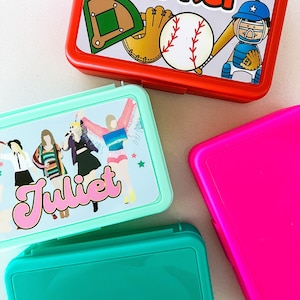 May include: Four rectangular plastic lunch boxes in red, mint green, teal, and hot pink. The red box features a baseball graphic with the name Parker. The mint green box displays a graphic of four figures and the name Juliet in pink script.