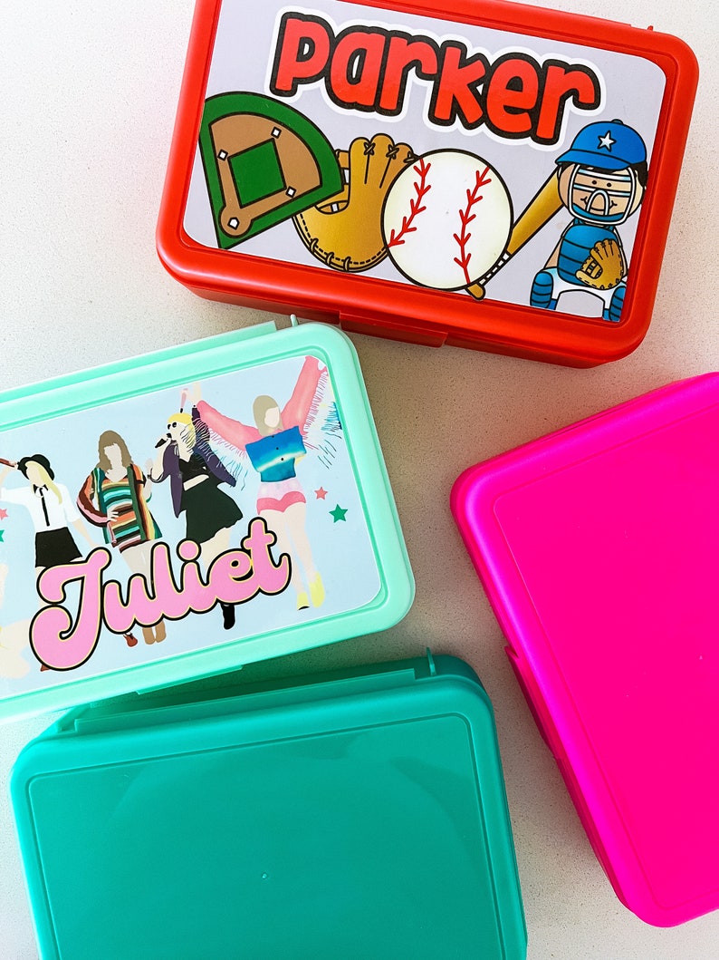 May include: Four rectangular plastic lunch boxes in red, mint green, teal, and hot pink. The red box features a baseball graphic with the name Parker. The mint green box displays a graphic of four figures and the name Juliet in pink script.