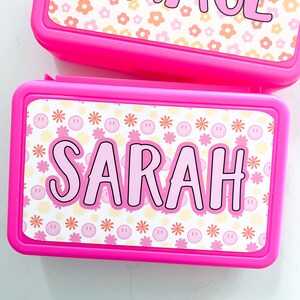 K&ouml;nnte beinhalten: Zwei rechteckige, leuchtend pinke Kunststoff-Lunchboxen mit personalisierten Aufklebern. Die obere Box zeigt den Namen GRACE in pinken Buchstaben auf einem pfirsichfarbenen und wei&szlig;en Blumenmuster. Die untere Box zeigt den Namen SARAH in pinken Buchstaben auf einem Muster aus pinken Smileys und orangen Blumen.