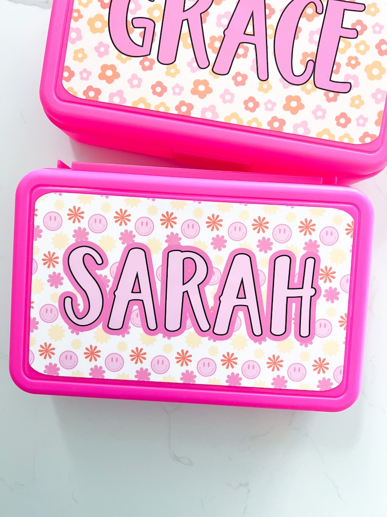 K&ouml;nnte beinhalten: Zwei rechteckige, leuchtend pinke Kunststoff-Lunchboxen mit personalisierten Aufklebern. Die obere Box zeigt den Namen GRACE in pinken Buchstaben auf einem pfirsichfarbenen und wei&szlig;en Blumenmuster. Die untere Box zeigt den Namen SARAH in pinken Buchstaben auf einem Muster aus pinken Smileys und orangen Blumen.