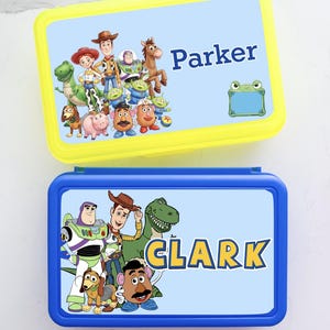 May include: Two rectangular plastic lunch boxes with light blue lids featuring Toy Story character graphics. The top yellow-rimmed box displays the name Parker in blue text. The bottom blue-rimmed box displays the name CLARK in yellow text.