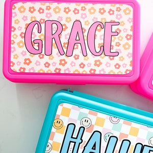 K&ouml;nnte beinhalten: Eine rechteckige, leuchtend pinke Brotdose mit Blumenmuster und dem Namen GRACE in pinken Buchstaben sowie eine rechteckige t&uuml;rkisfarbene Brotdose mit kariertem Smiley-Muster und dem Namen HALLIE in blauen Buchstaben auf den Deckeln.
