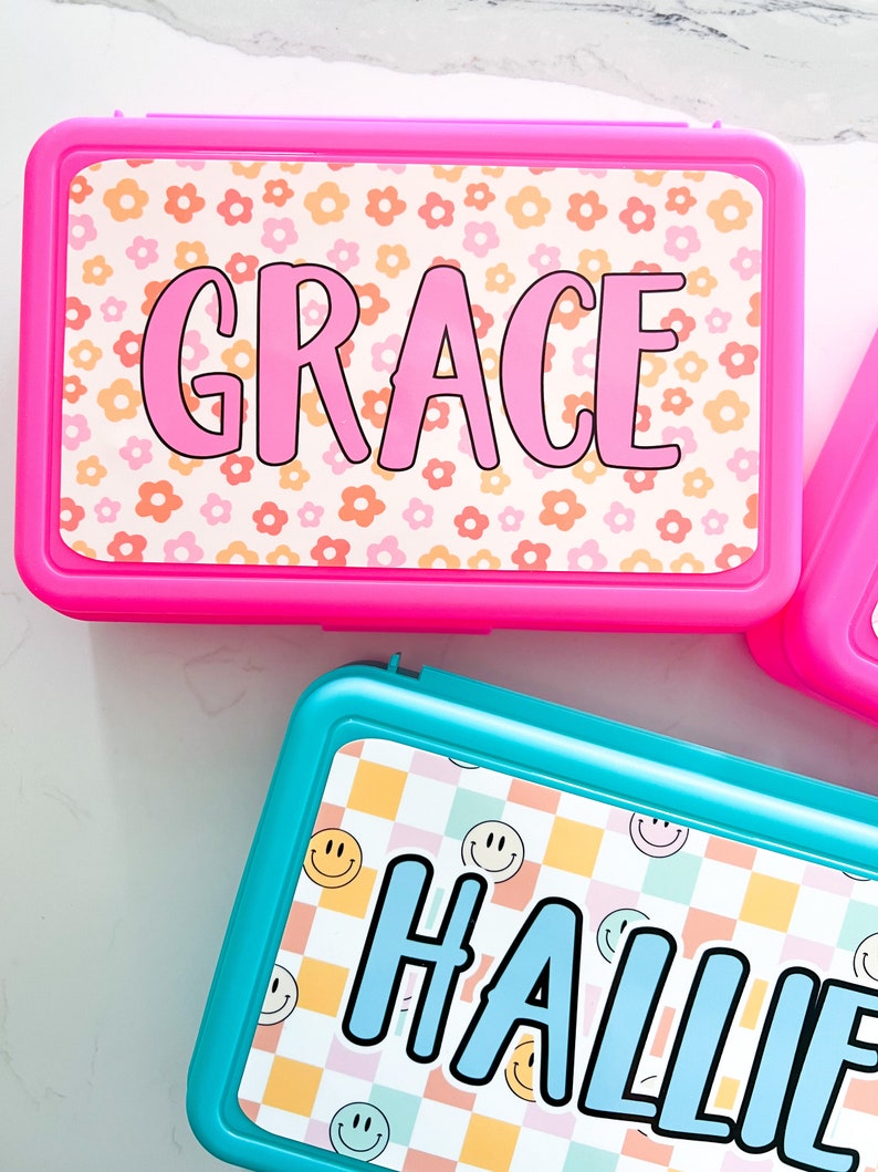 K&ouml;nnte beinhalten: Eine rechteckige, leuchtend pinke Brotdose mit Blumenmuster und dem Namen GRACE in pinken Buchstaben sowie eine rechteckige t&uuml;rkisfarbene Brotdose mit kariertem Smiley-Muster und dem Namen HALLIE in blauen Buchstaben auf den Deckeln.