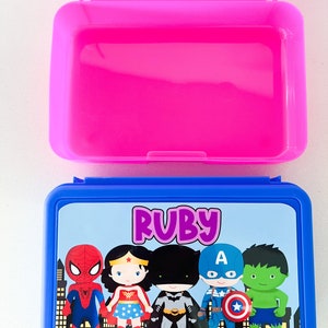 May include: A rectangular blue plastic lunch box with a hinged lid featuring a graphic print of five cartoon superheroes and the name Ruby in purple text. The interior of the lunch box is bright pink and is shown open with the lid flipped upward.