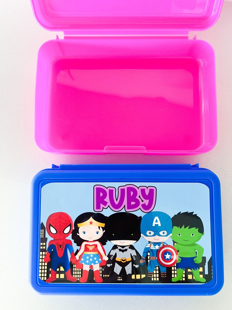 May include: A rectangular blue plastic lunch box with a hinged lid featuring a graphic print of five cartoon superheroes and the name Ruby in purple text. The interior of the lunch box is bright pink and is shown open with the lid flipped upward.