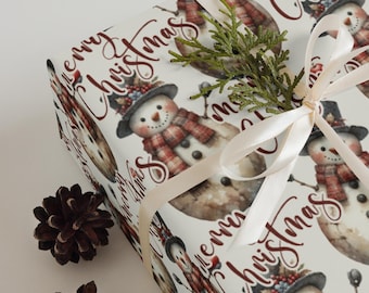 Op de afbeelding: Kerstcadeaupapier met een herhalend sneeuwpopontwerp, compleet met hoeden en tartan sjaals. De woorden "Merry Christmas" zijn geschreven in een cursief schrift. Een crèmekleurig lint en een takje groen sieren het verpakte cadeau.