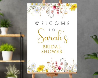 May include: A welcome sign for Sarah's bridal shower, displayed on a wooden easel. The sign features floral watercolor artwork with daisies, yellow flowers, and other wildflowers. The text reads "Welcome to Sarah's Bridal Shower."