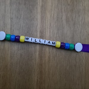 May include: A personalized keychain with the name "WILLIAM" spelled out in white letter beads. The beads are interspersed with colorful beads in green, blue, purple, and yellow. The keychain has a purple ribbon and a silver key ring.