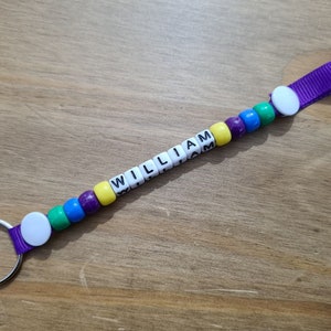 May include: A handmade keychain with the name "WILLIAM" spelled out in white letter beads. The beads are interspersed with colorful beads in purple, yellow, green, and blue. A purple ribbon and a white button secure the beads to the keyring.