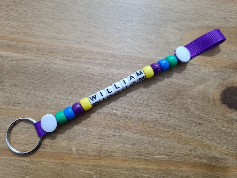 May include: A handmade keychain with the name "WILLIAM" spelled out in white letter beads. The beads are interspersed with colorful beads in purple, yellow, green, and blue. A purple ribbon and a white button secure the beads to the keyring.