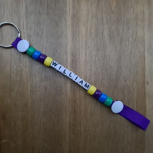 May include: A handmade keychain with the name "WILLIAM" spelled out in white letter beads. The keychain features colorful beads in green, blue, purple, and yellow. A purple ribbon and a silver key ring complete the design.