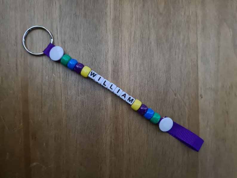 May include: A handmade keychain with the name "WILLIAM" spelled out in white letter beads. The keychain features colorful beads in green, blue, purple, and yellow. A purple ribbon and a silver key ring complete the design.