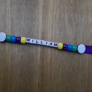 May include: A personalized keychain with the name "WILLIAM" spelled out in white letter beads. The beads are interspersed with colorful beads in green, blue, purple, and yellow. The keychain has a purple ribbon and a silver key ring.