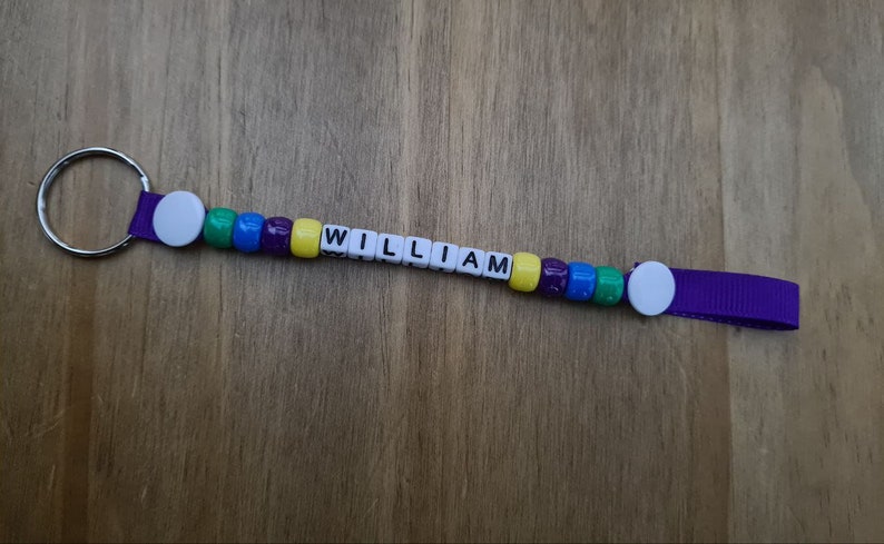 May include: A personalized keychain with the name "WILLIAM" spelled out in white letter beads. The beads are interspersed with colorful beads in green, blue, purple, and yellow. The keychain has a purple ribbon and a silver key ring.