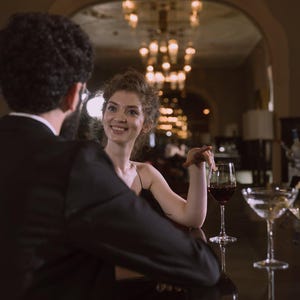 K&ouml;nnte beinhalten: Ein Mann und eine Frau sitzen an einer Bar. Die Frau l&auml;chelt und h&auml;lt ein Glas Rotwein in der Hand. Der Mann tr&auml;gt einen schwarzen Anzug und die Frau ein schwarzes Kleid.