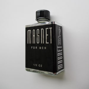 K&ouml;nnte beinhalten: Eine schwarze Glasflasche mit einem silbernen Etikett, auf dem "MAGNET FOR MEN 1/2 OZ" steht. Die Flasche hat einen schwarzen Verschluss und steht auf einem wei&szlig;en Hintergrund.