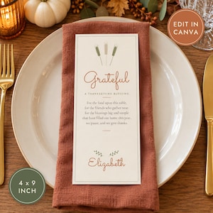 May include: A rectangular paper place card measuring 4 by 9 inches, featuring the text Grateful A Thanksgiving Blessing, a prayer, and the name Elizabeth in rust-colored script, centered on a rust-colored linen napkin atop a white plate.